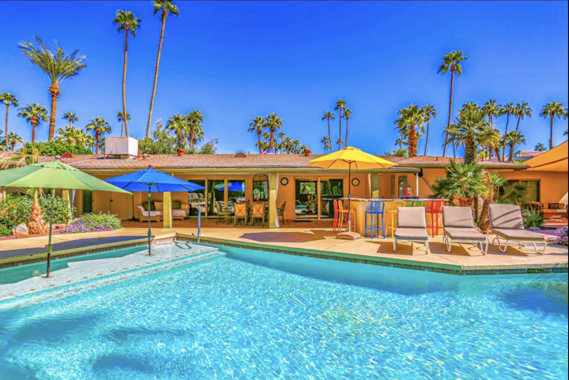 Palm Paradise - Palm Springs, CA Luxurious Palm Springs vacation home with a pool, colorful umbrellas, palm trees, and outdoor seating in a sunny desert setting.