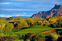 Scenic view of a golf course with lush green fairways, surrounded by desert vegetation and rugged mountains under a partly cloudy sky.