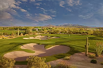 A scenic golf course with sand traps, cacti, and lush green fairways under a partly cloudy sky, with mountains in the background.