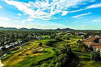 A scenic view of Stonecreek Golf Club with lush green fairways, trees, residential buildings, and mountains under a bright blue sky.