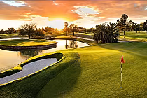 Beautiful golf course at sunset with lush green grass, water hazards, palm trees, and a red flag on the putting green.