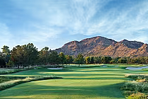 A scenic golf course with lush green fairways, sand bunkers, trees, and mountains in the background under a blue sky.