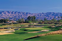 Scenic view of La Quinta PGA WEST Pete Dye Dunes golf course with lush green fairways, sand traps, water hazards, palm trees, and mountains in the background.