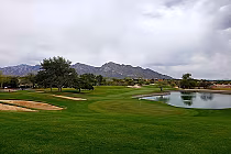 Scenic desert golf course with lush green fairways, sand bunkers, a water hazard, and mountains in the background under cloudy skies.