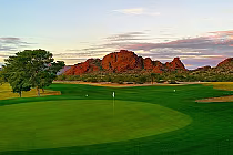 Scenic view of a golf course with a green, flag, trees, and red rock formations in the background at sunset in Phoenix, AZ.