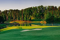 Scenic view of a golf course at sunrise with lush green fairways, sand bunkers, a water hazard, and a dense forest in the background.