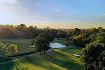 Scenic view of Highland Park Golf Course with lush green fairways, trees, a pond, and a city skyline in the distance during sunset.