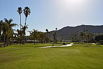 View of Indian Canyons Golf Resort with lush green fairways, tall palm trees, mountains in the background, and clear blue sky.