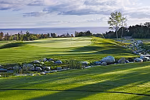 Scenic view of Lora Bay Golf Club with lush green fairways, a water feature with rocks, and a distant view of the ocean under a partly cloudy sky.