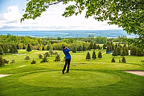 A golfer swings a club on a lush green golf course with trees, scenic views, and a distant body of water under a partly cloudy sky.