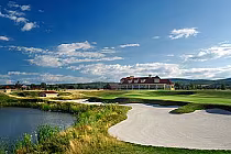 Scenic view of Batteaux Creek Golf Club with a large house, sand traps, water hazard, and lush green fairways under a blue sky.