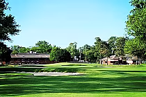 A lush green golf course with sand traps, surrounded by trees and residential buildings, under a clear blue sky.
