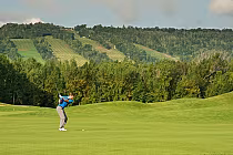A golfer in a blue shirt and gray pants swings a club on a lush green golf course with a wooded area and hills in the background.