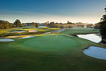 Scenic view of a golf course with a green, sand bunkers, water hazards, trees, and a sunrise or sunset sky.