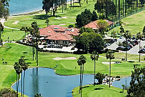 Aerial view of Coronado Golf Course with lush green fairways, water hazards, a clubhouse, and parking lot near San Diego harbor.
