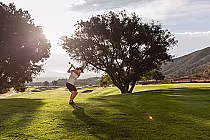 A golfer swings a club on a scenic, lush golf course with trees, hills, and mountains in the background during sunset.