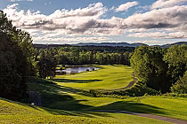 Scenic view of La Bête golf course at Mont Tremblant with lush green fairways, trees, a pond, and distant mountains under a partly cloudy sky.