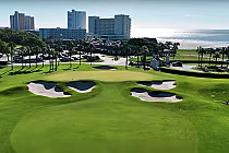 Aerial view of Dunes Golf & Beach Club with lush green fairways, sand bunkers, palm trees, beachfront, and high-rise buildings in the background.