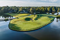 A scenic golf course with a green, sand bunkers, water hazards, surrounded by trees and a few houses, under a partly cloudy sky.