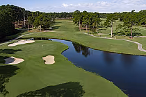 A scenic golf course with sand bunkers, a water hazard, lush green fairways, and tall trees under a partly cloudy sky.