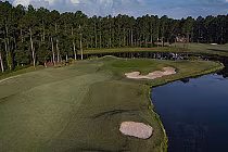 A scenic golf course with lush green fairways, sand bunkers, surrounded by tall trees and a calm water hazard.