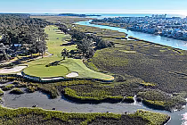 Aerial view of a golf course with lush green fairways, sand traps, trees, and water bodies, overlooking a coastal town and ocean.