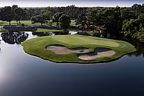 Aerial view of a golf course island green with sand bunkers, surrounded by water, lush trees, and a clear sky at Tradition Golf Club.