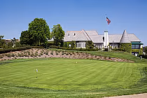 A lush green golf course with a clubhouse in the background, featuring a flagpole with an American flag and well-maintained landscaping.