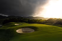 A lush green golf course with sand bunkers, surrounded by trees and mountains under a dramatic cloudy sky with sunlight breaking through.