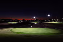 Nighttime view of a golf course with illuminated putting green, rolling hills, and bright lights under a dark sky with a hint of sunset.