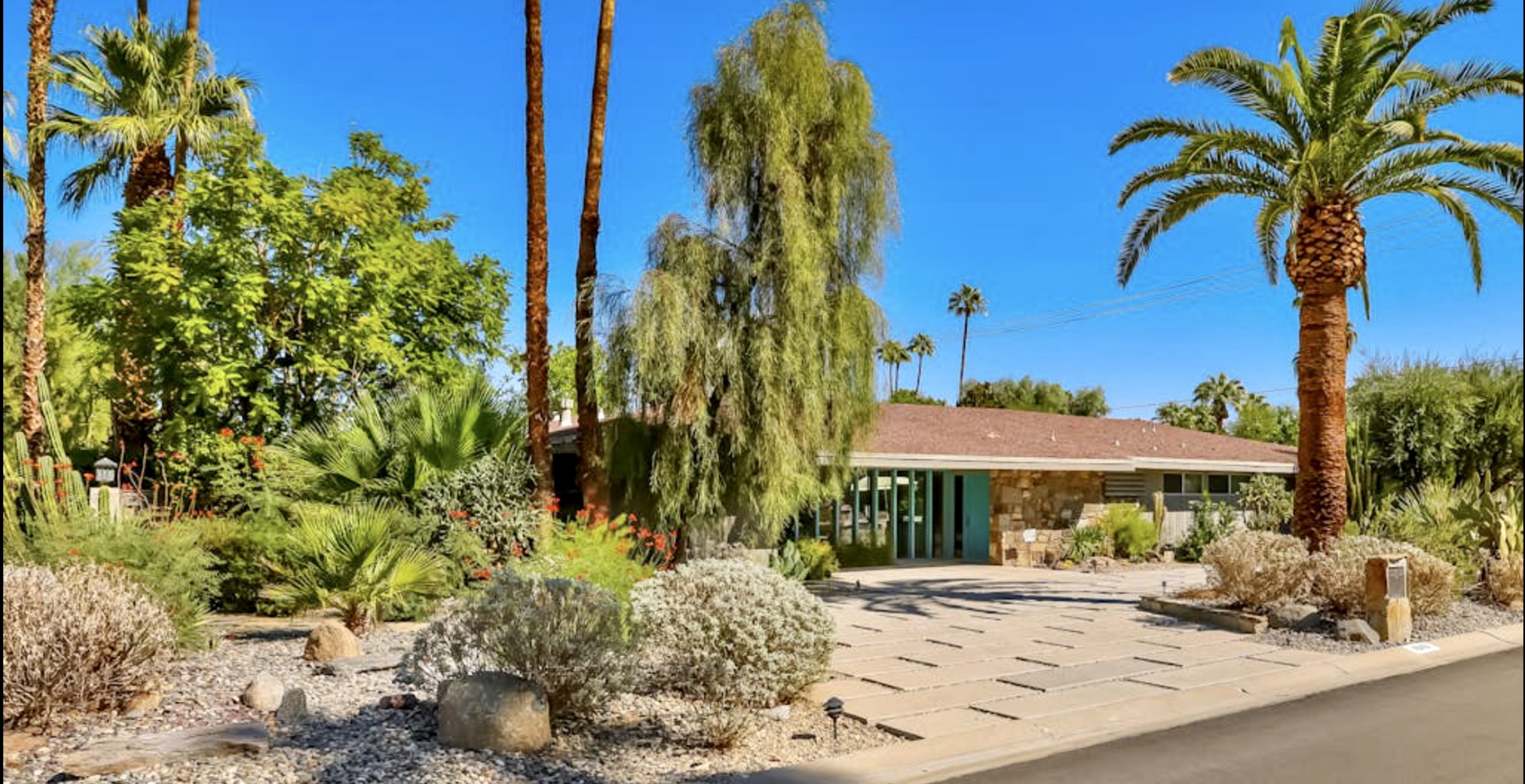 Palm Springs Perfection A modern Palm Springs vacation home with a stone exterior, surrounded by desert plants, palm trees, and a clear blue sky.