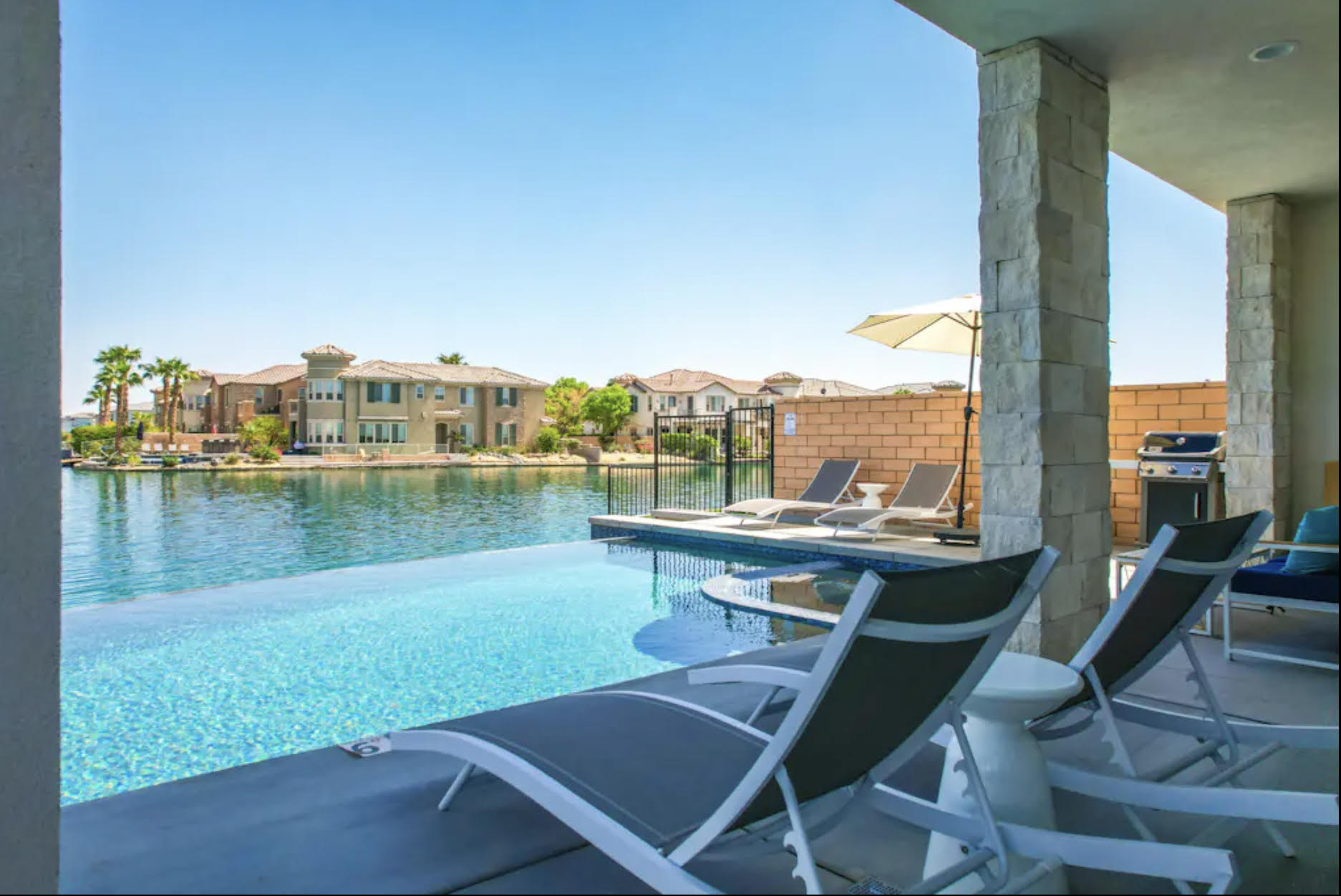 Lakeside Infinity - House Indio Luxury lakeside pool area with lounge chairs, umbrella, and scenic view of waterfront homes in Indio, California.