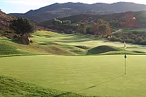 Scenic view of a golf course with a putting green, flag, rolling hills, trees, and mountains in the background at Steele Canyon Golf Club.
