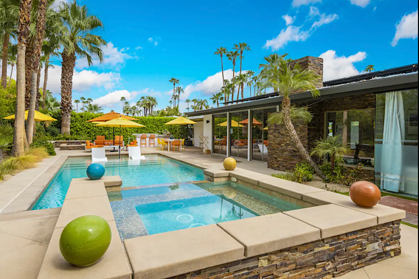 The High Road - 3400 - Palm Springs, CA Luxury backyard with a pool, hot tub, colorful spheres, palm trees, outdoor seating, umbrellas, and scenic mountain views in Palm Springs.