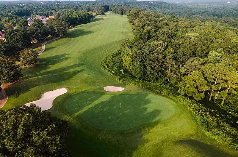 Aerial view of a lush golf course in Alabama with green fairways, sand bunkers, surrounded by dense trees and a distant landscape.