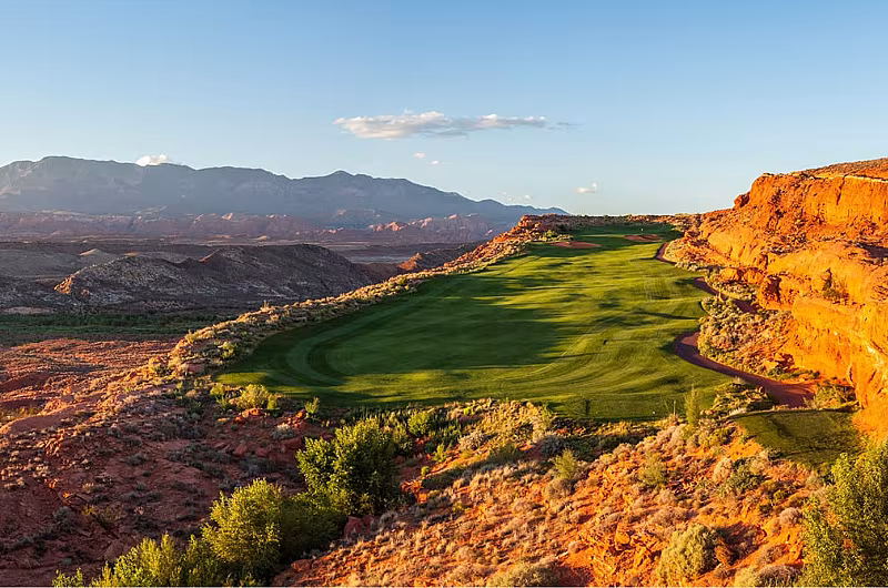 Scenic view of a golf course nestled in a desert landscape with red rock formations and distant mountains under a clear sky.