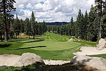 A scenic golf course with lush green fairways, surrounded by tall pine trees and large rocks, under a partly cloudy sky.