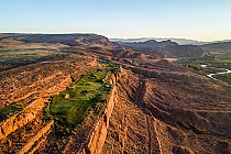Aerial view of Sand Hollow Golf Course with lush green fairways on red rock cliffs in Utah's desert landscape at sunset.