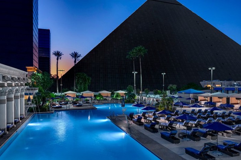 Luxor Hotel Luxor Hotel in Las Vegas with a large illuminated pool area, surrounded by lounge chairs and umbrellas, at dusk.