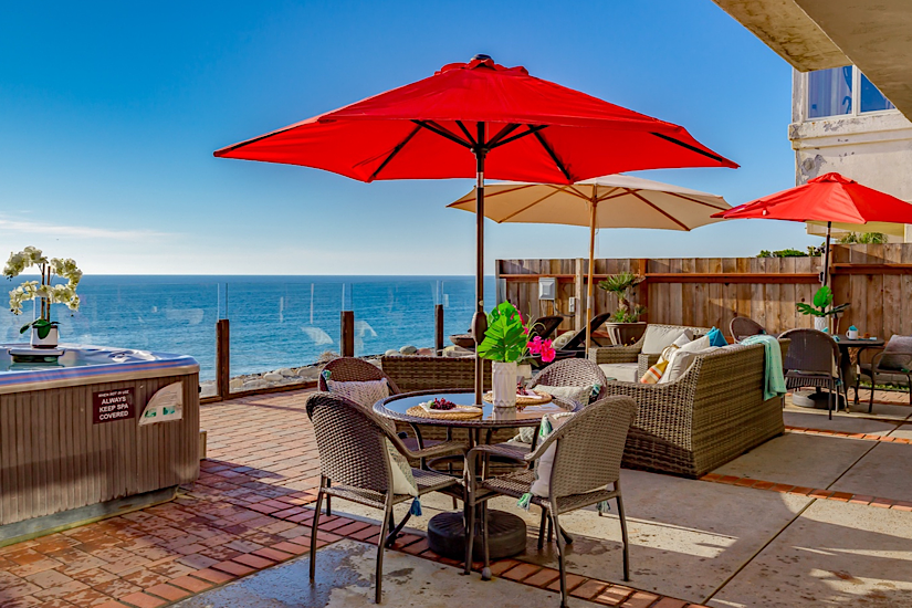 Outdoor patio with wicker furniture, umbrellas, and a hot tub overlooking the Pacific Ocean from a luxurious home on a bluff.