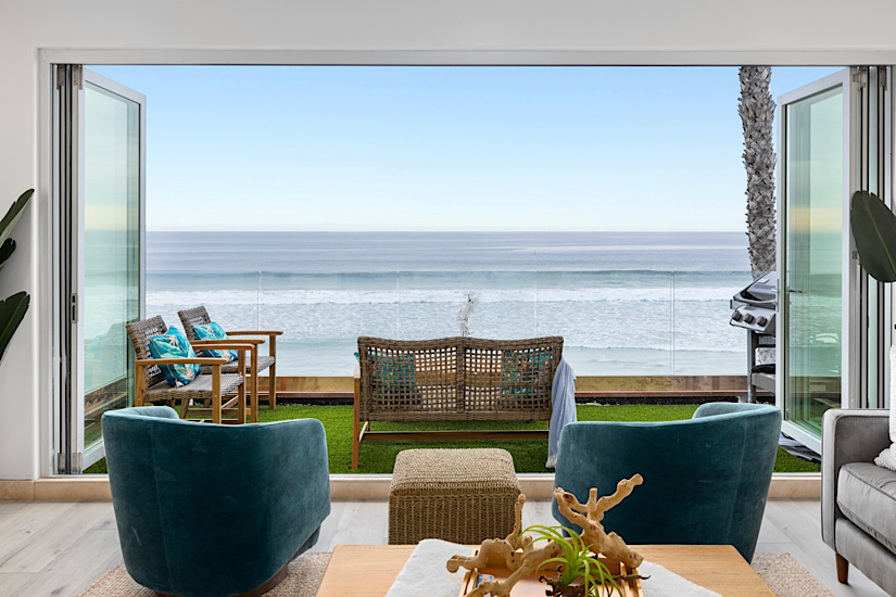 View from inside a beachfront rental living room looking out to a rooftop deck with ocean view, outdoor seating, hot tub, and BBQ.