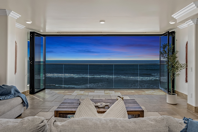 Interior view of a living room with open glass doors leading to a balcony overlooking the ocean at sunset, with a cozy sofa and potted plant.