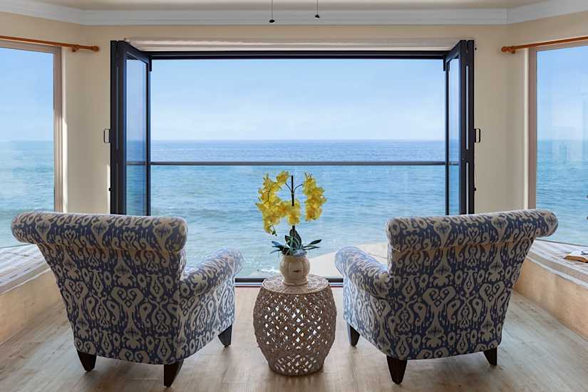Interior of a beachfront vacation home with two patterned armchairs facing large open windows overlooking the ocean.