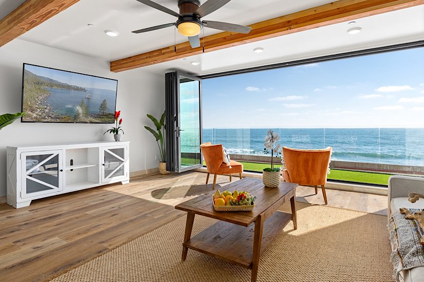 Bright living room with large glass doors opening to ocean view, featuring cozy chairs, wooden table, and modern decor.