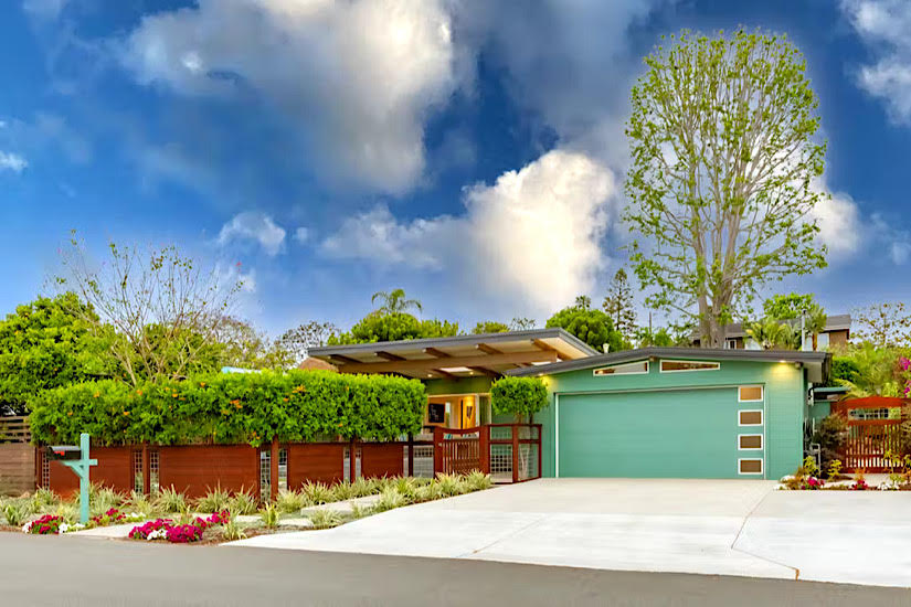 A modern, colorful cottage with a green garage door, surrounded by lush greenery and vibrant flowers under a partly cloudy sky.