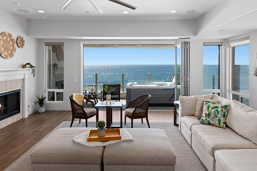 Bright living room with large windows and ocean view, featuring a white sofa, fireplace, dining area, and a hot tub on the balcony.