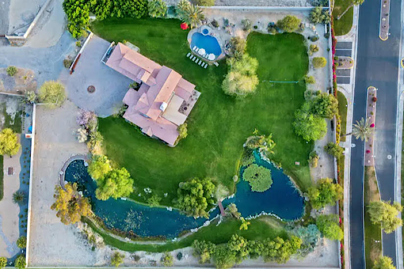 The Coachella Estate Aerial view of The Coachella Estate, a luxury resort with lush green lawns, a swimming pool, ponds, and surrounding trees.