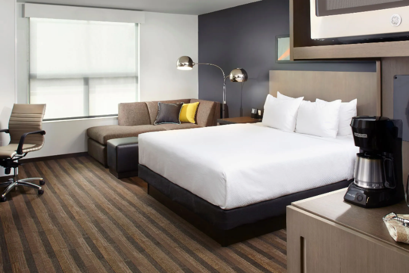 Modern hotel room at Hyatt House Lakewood with a large bed, sofa, desk, and window with natural light, overlooking Denver.