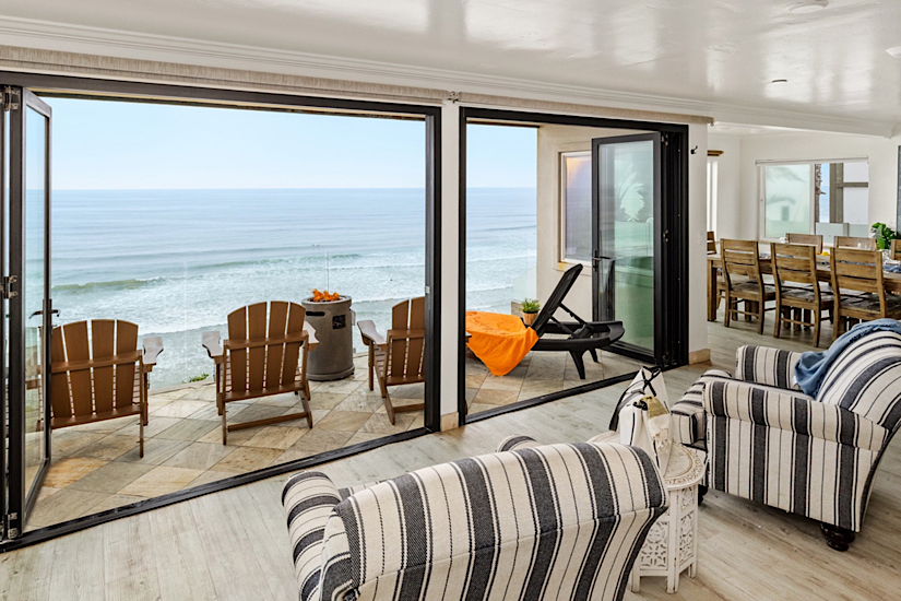 Interior of a beachfront condo with large glass doors opening to a balcony overlooking the Pacific Ocean, with chairs and a lounge chair.