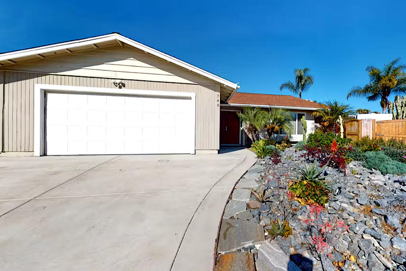 Sunlit house with a white garage door, surrounded by a colorful garden and palm trees under a clear blue sky in Vista, CA.